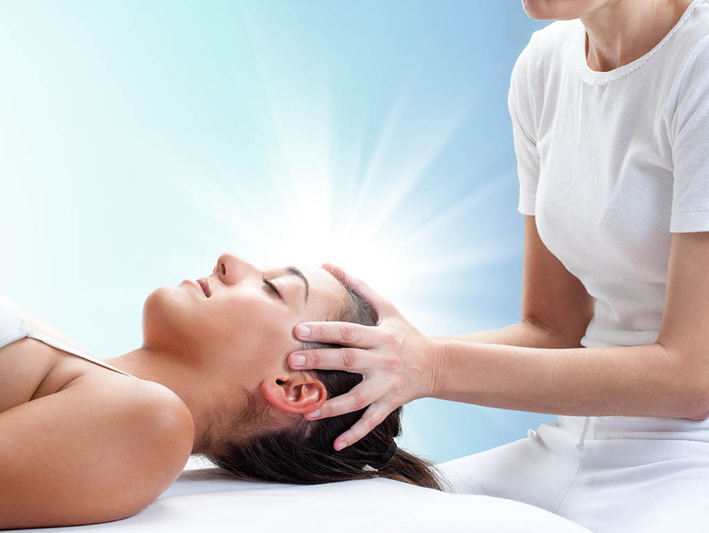 A woman receiving a Reiki treatment from a practitioner, lying down with her eyes closed.